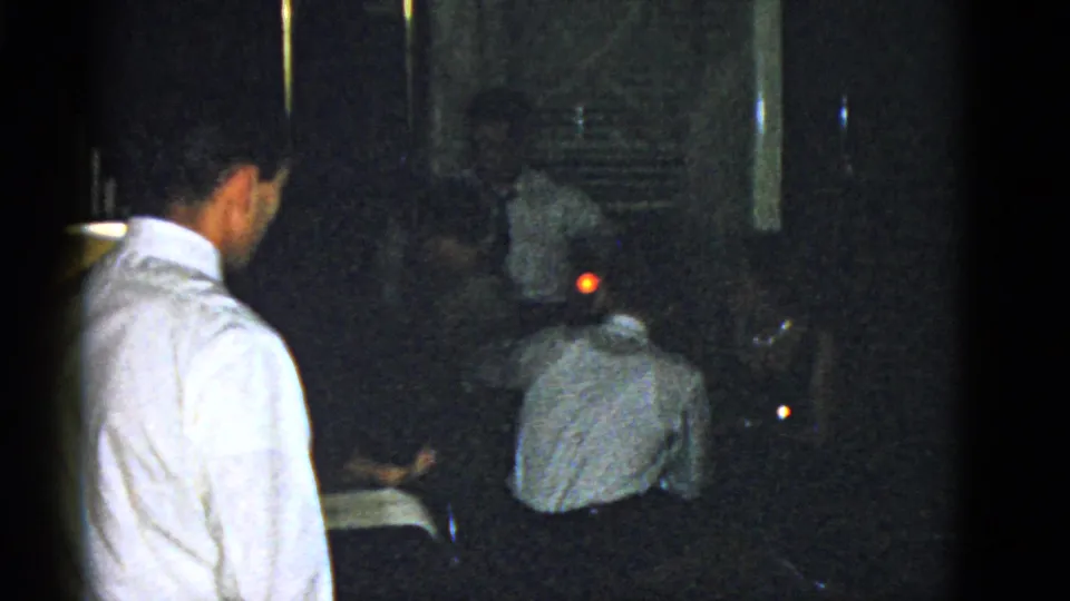 A dark 1951 Boston home-movie frame showing adults gathered indoors, with a standing man and a red point of light behind seated figures.