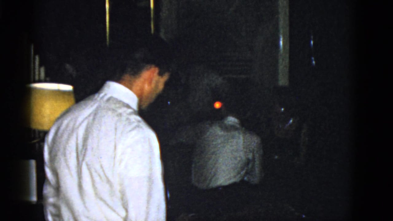 Man in a white dress shirt stands in a dark room while another man sits ahead of him.