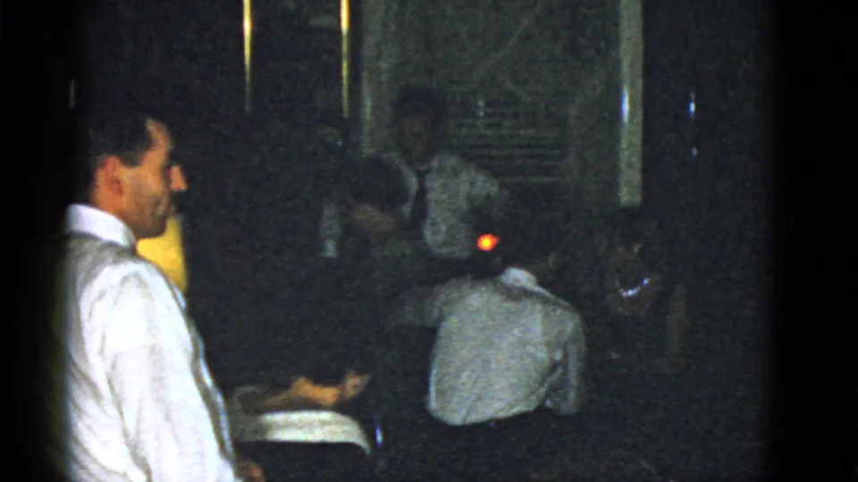 Side view of a man in a white shirt during a 1951 Boston indoor gathering, with seated figures and a lamp nearby.