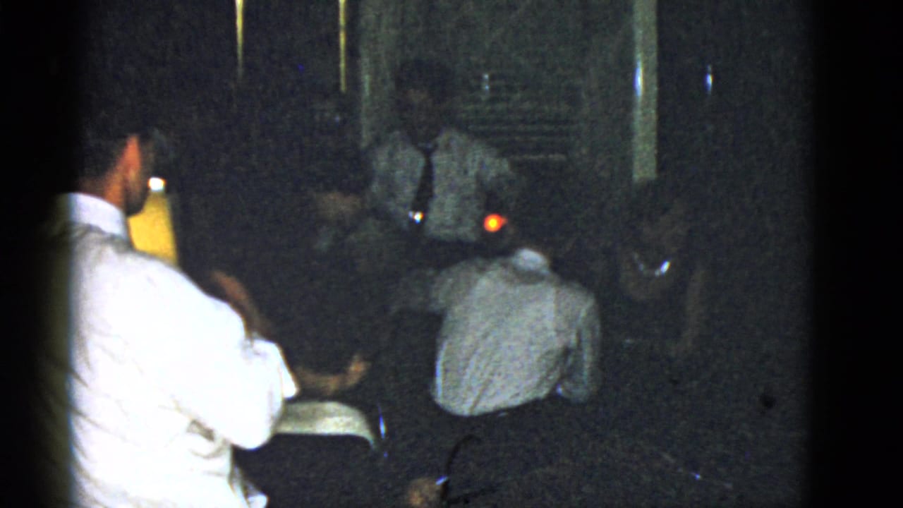 Adults gathered in a dim room, with a man in a white shirt near the foreground and a lamp glowing in the background.