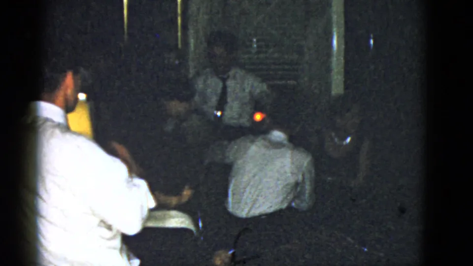1951 Boston home-movie frame showing adults at a dark indoor party, including men in white shirts and a small glowing lamp.