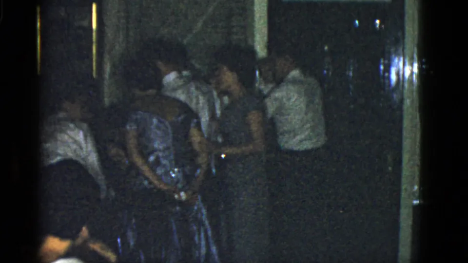 1951 Boston home-movie frame of a crowded indoor party, centered on adults standing near a woman in a blue dress.