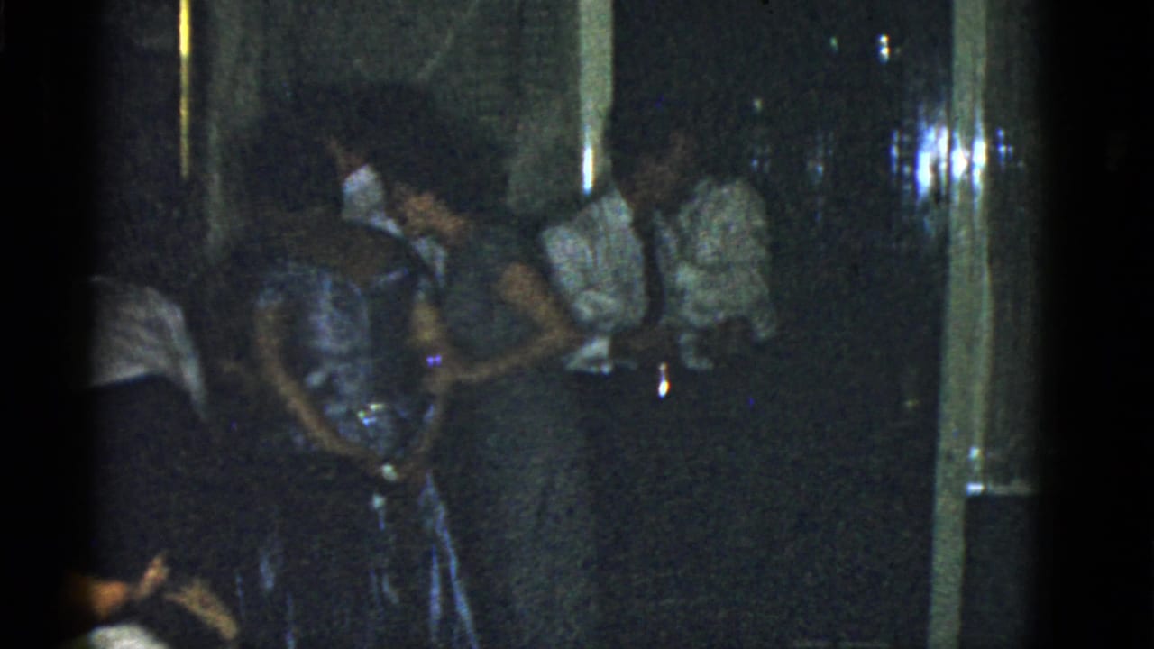 1951 Boston home-movie frame showing adults in dresses, shirts, and ties gathered in a dark indoor party space.