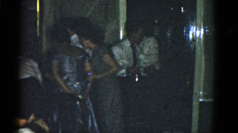 1951 Boston home-movie frame showing adults in dresses, shirts, and ties gathered in a dark indoor party space.