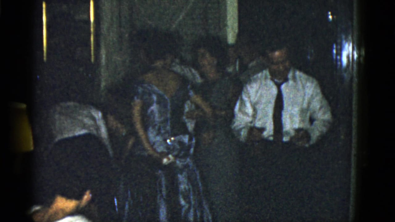 Adults in formal clothing gather in a dim room, with a man in a white shirt holding drinks.