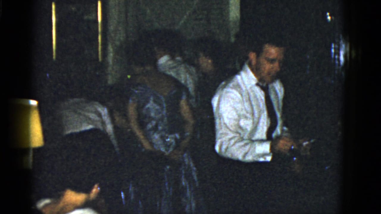 A man in a white shirt and tie stands beside guests in dresses in a dim interior.