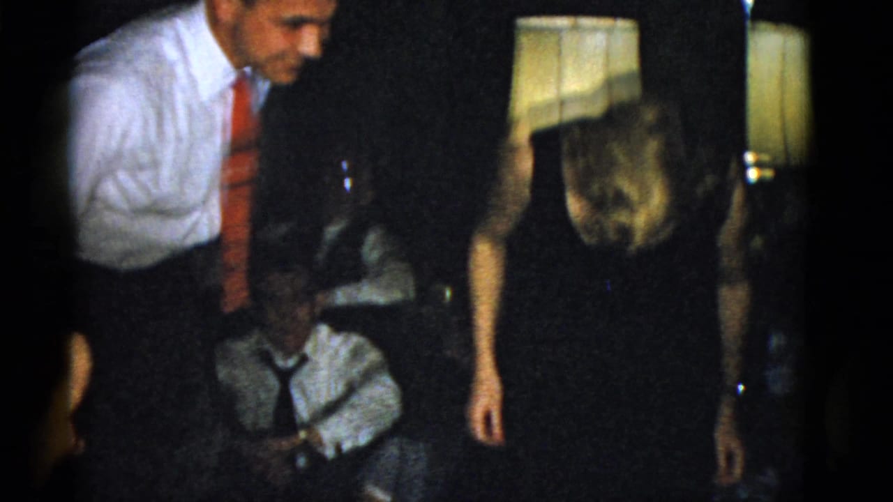 Man in white shirt and red tie stands near seated guests in a dim interior.