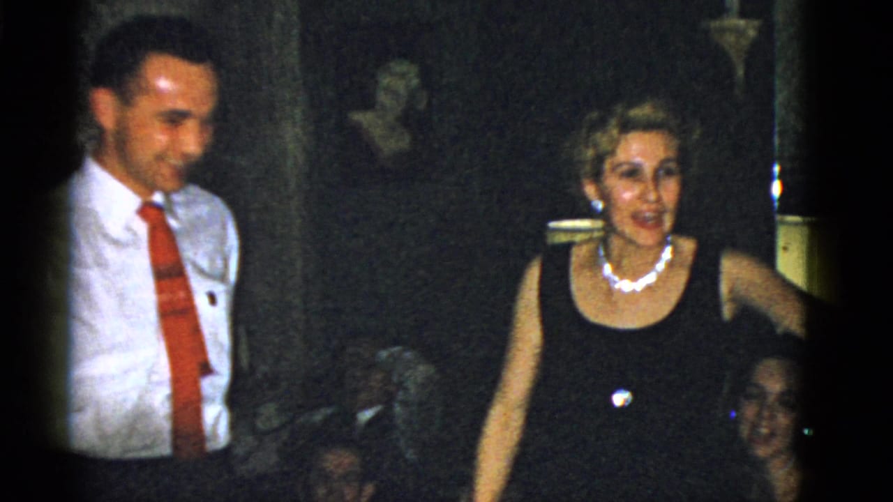 Man in red tie stands beside a woman in a black dress and pearl necklace.