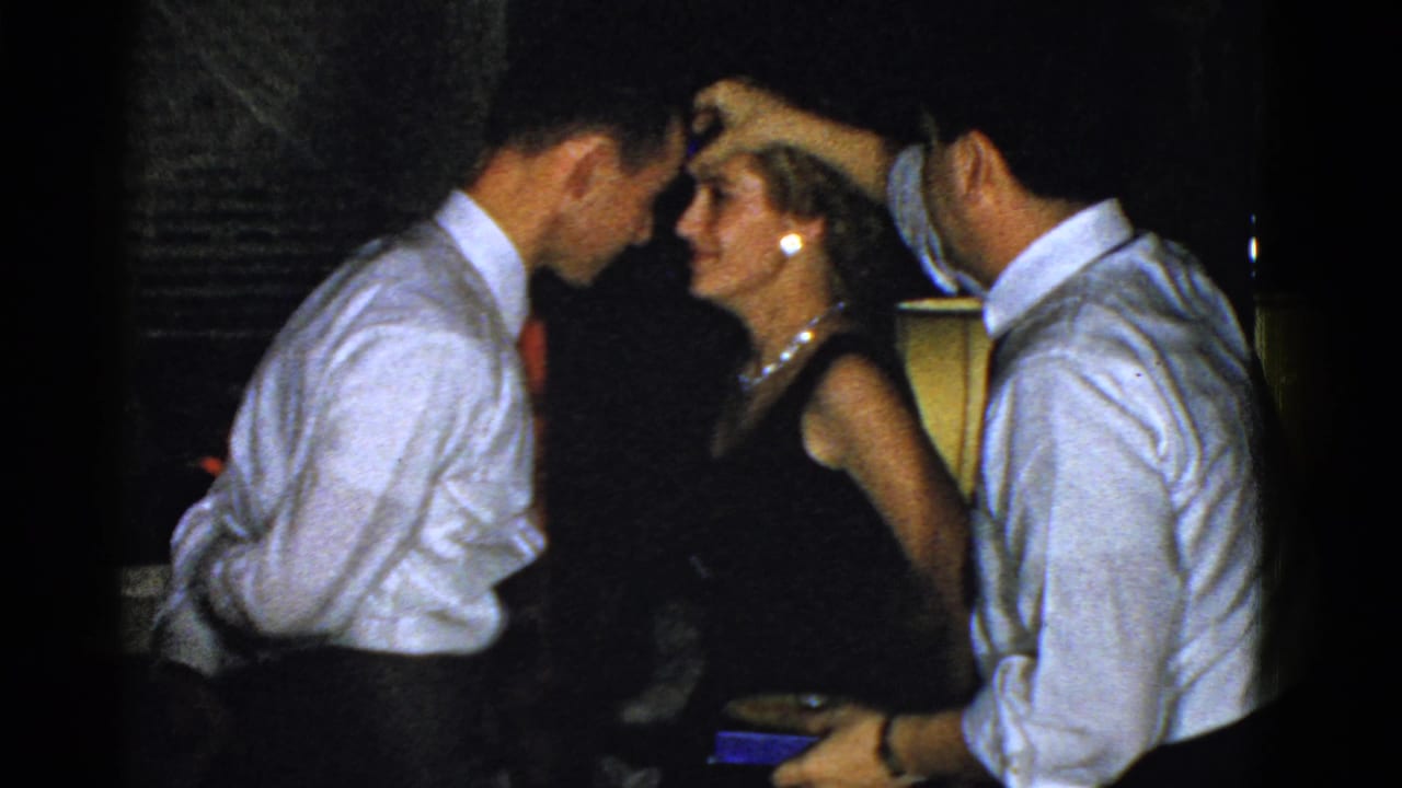 Woman in a dark dress stands between two men in white shirts in a dim interior.