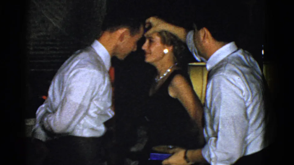 In a 1951 Boston home-movie frame, a woman in formal dress stands close to two men in white shirts inside a dim room.