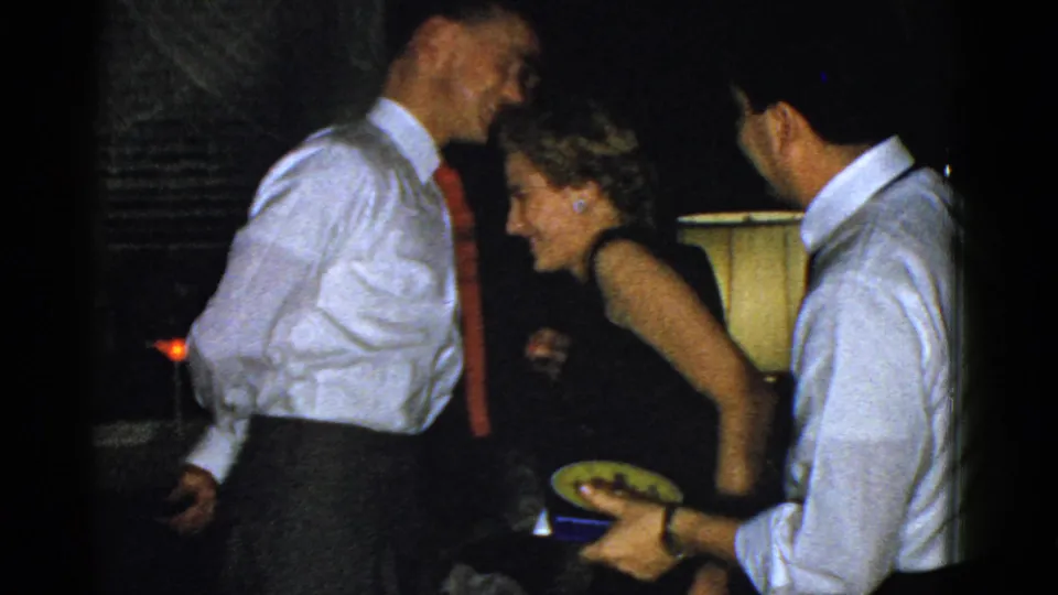 At the Boston gathering, the man in the red tie and a woman laugh while another man in a white shirt holds a plate near a lit lamp.