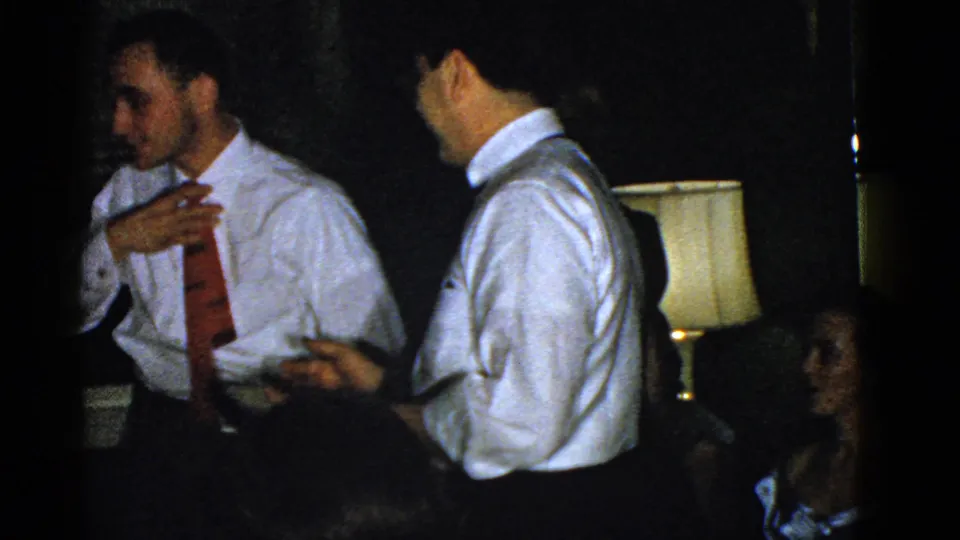 The man in the red tie gestures near his chest as another man in a white shirt faces him with a plate in the dim Boston interior.