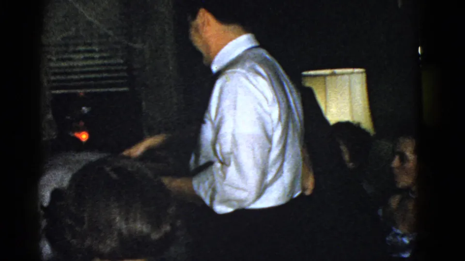 A man in a white shirt and suspenders stands near a glowing lamp, with seated guests partly visible in the 1951 Boston home-movie scene.