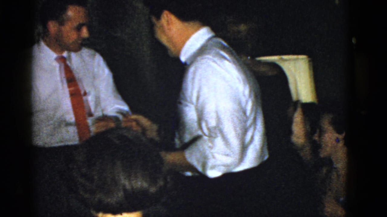 Two men in white shirts and ties shake hands in a dim room with seated guests nearby.