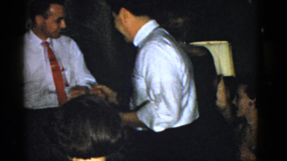In a 1951 Boston home-movie frame, two men in dress shirts and ties shake hands during a dim indoor gathering.