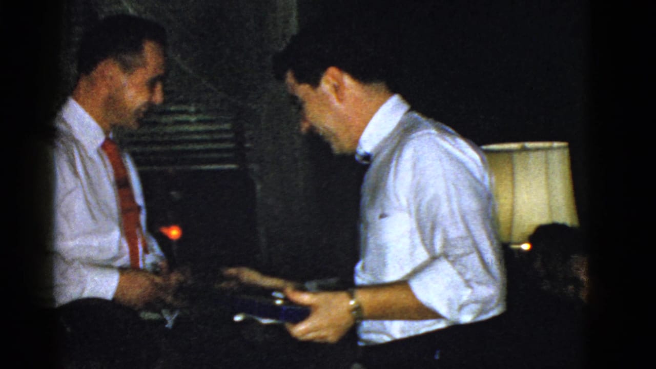 A man with a dark tie holds a small blue box while facing a man with a red tie.