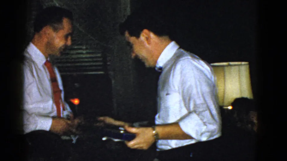 A 1951 Boston indoor frame shows a man holding a small blue box as he faces another man in a red tie.