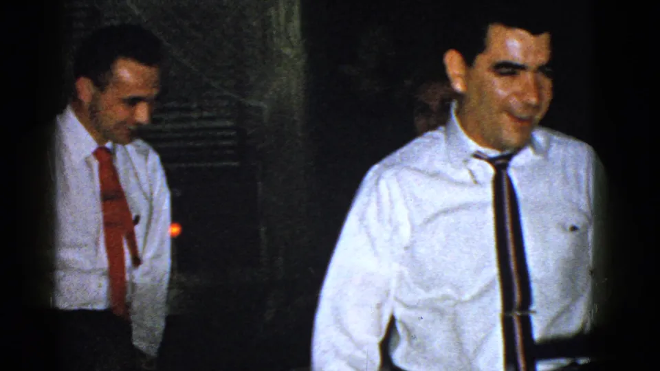 In this 1951 Boston home-movie frame, two men in white shirts and ties stand near guests in a low-lit interior.