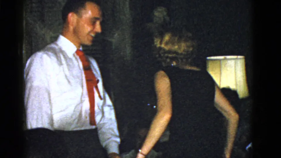 A 1951 Boston home-movie frame shows a man in a red tie near a woman in a black dress and a lit lamp.