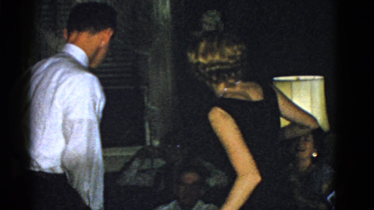 Man in a white shirt and woman in a black dress stand in a dim interior near seated guests.