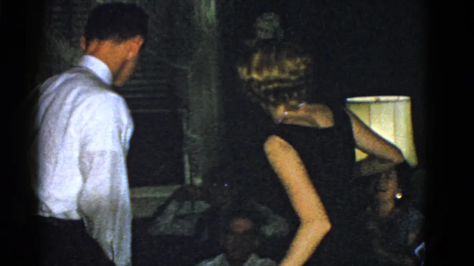 In 1951 Boston home-movie footage, adults in formal clothing gather in a dark private interior with a lamp and seated guests nearby.