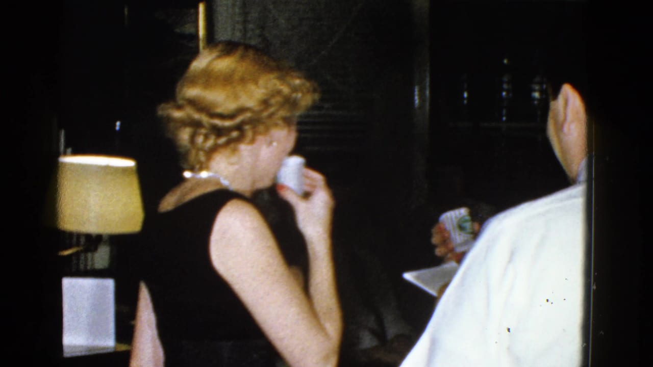 Woman in a black dress lifts a small cup while a man in a white shirt stands at the right edge.