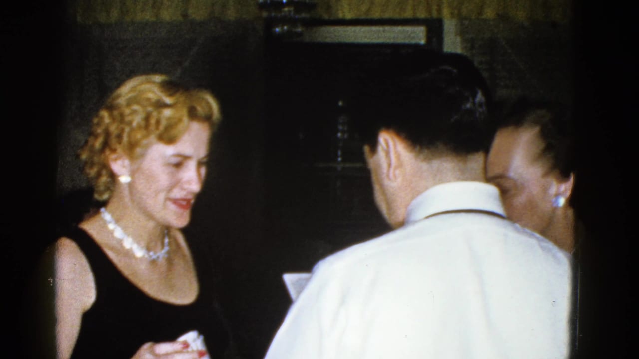 Blonde woman in a black dress smiles while standing with adults in a dim interior.