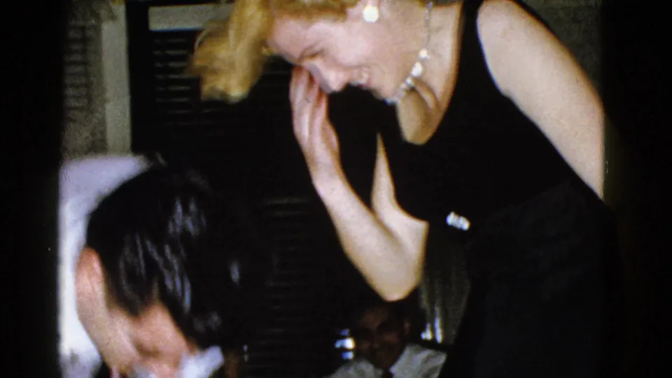 A blonde woman in a black dress leans forward with a broad smile near seated adults in 1951 Boston home-movie footage.