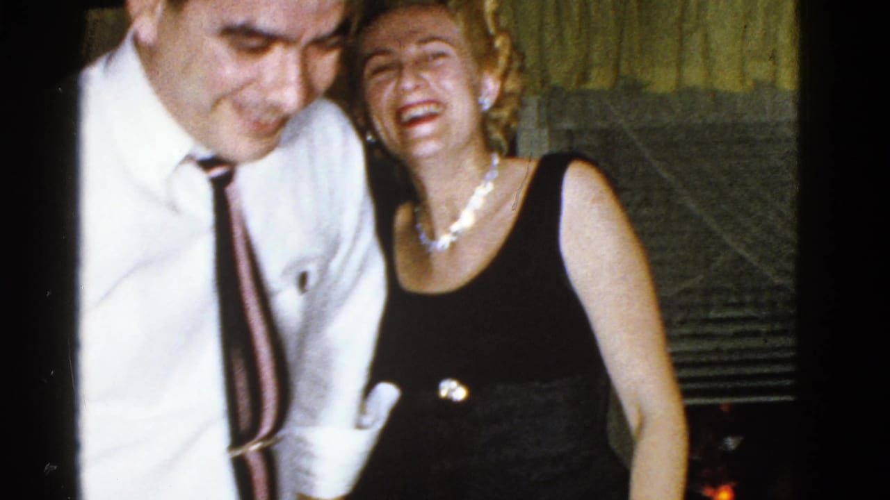 Man in a white shirt and tie laughs beside a woman in a black dress indoors.