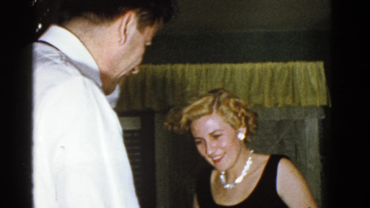 A man in a white shirt faces a smiling woman wearing a black sleeveless dress and pearl-style jewelry in a Boston interior.
