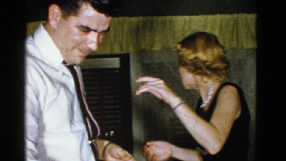The woman turns and raises one hand as the man stands nearby, with blinds and curtains visible in the 1951 home-movie frame.