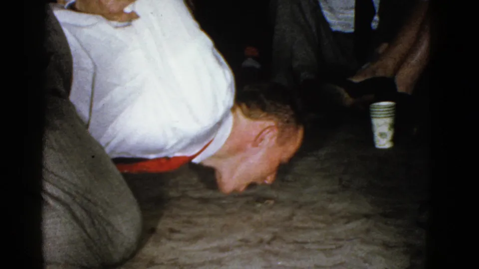 A man in a white shirt and red belt holds an upside-down pose on the carpet during the Boston workplace break, with seated coworkers and cups nearby.