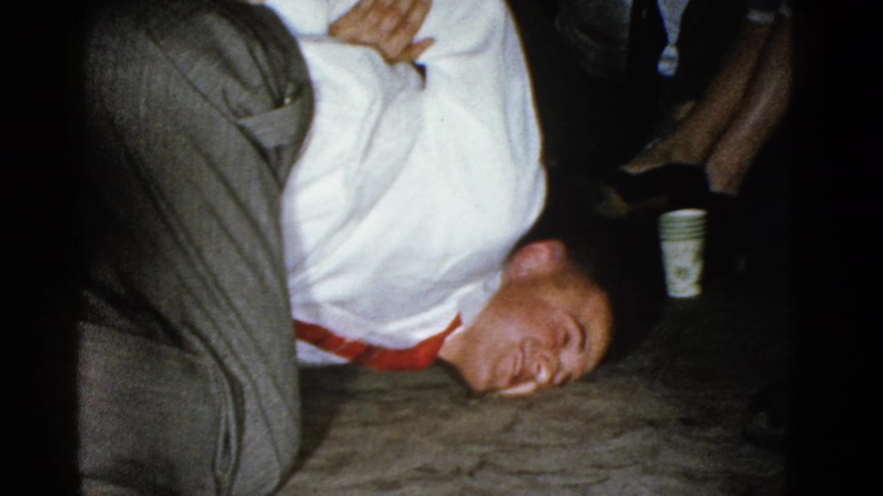 Man in a white shirt and red tie lies sideways on a floor near seated adults.