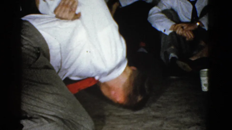 The home-movie frame shows the man close to the floor in a dim Boston interior, surrounded by formally dressed seated adults.