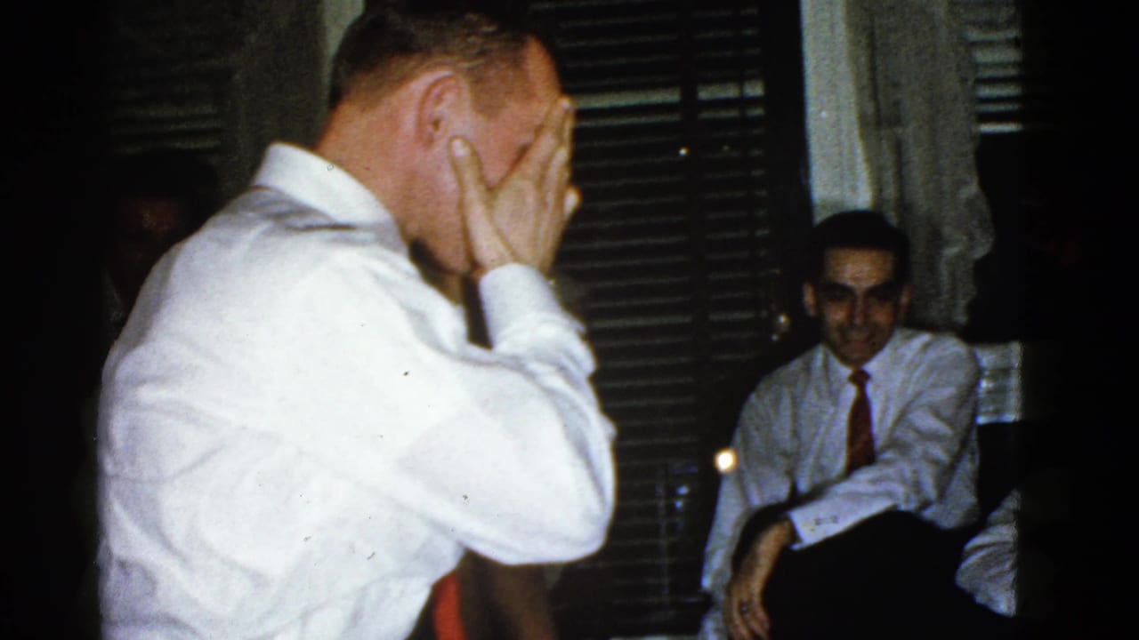 In a 1951 Boston home-movie frame, a man in a white shirt and tie acts during charades as guests sit near window blinds.