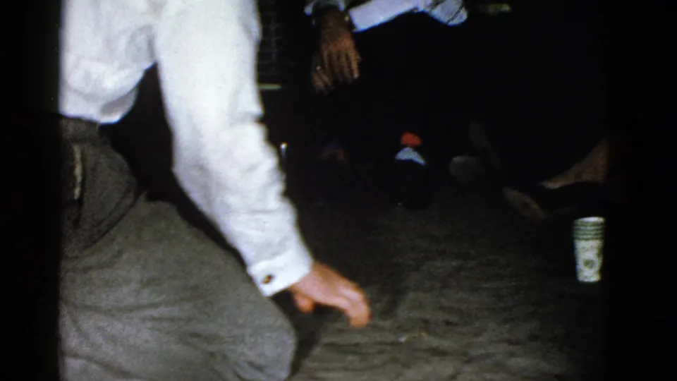 A man in a white shirt and tie crouches on the carpet during the party game, with dimly lit guests seated along the wall.