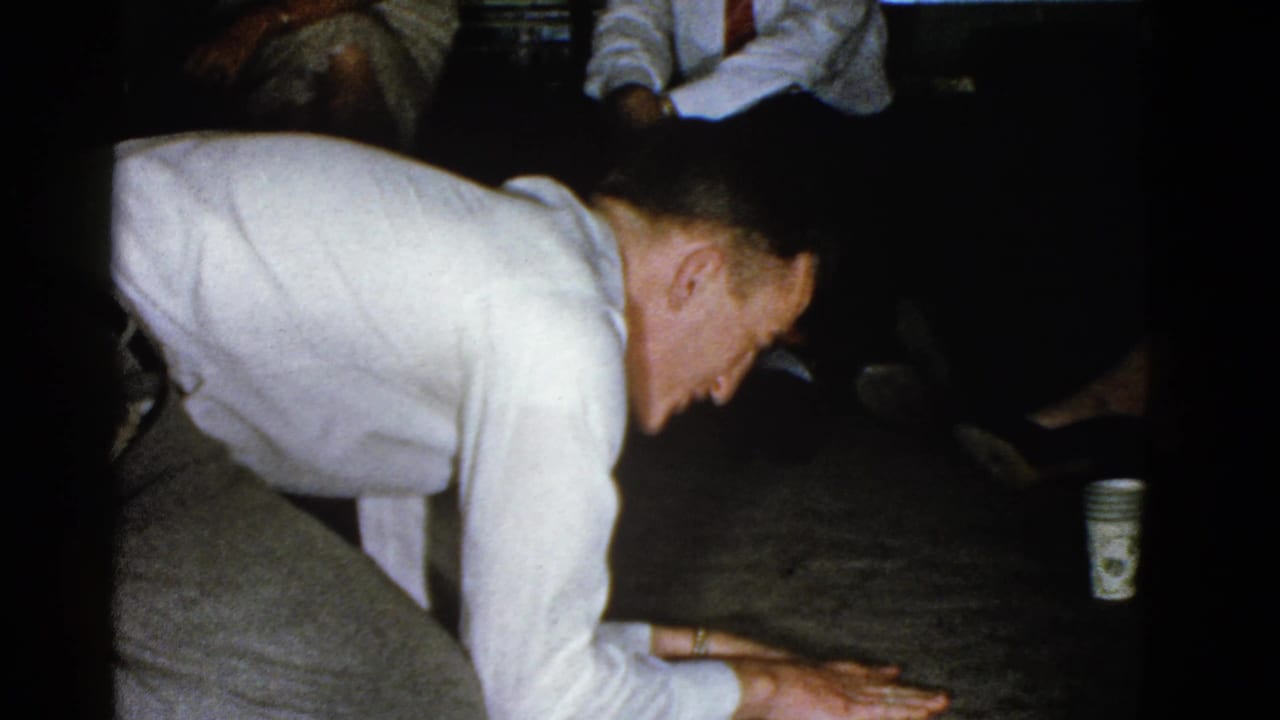 Man in a white shirt kneels on a carpeted floor while people sit behind him.