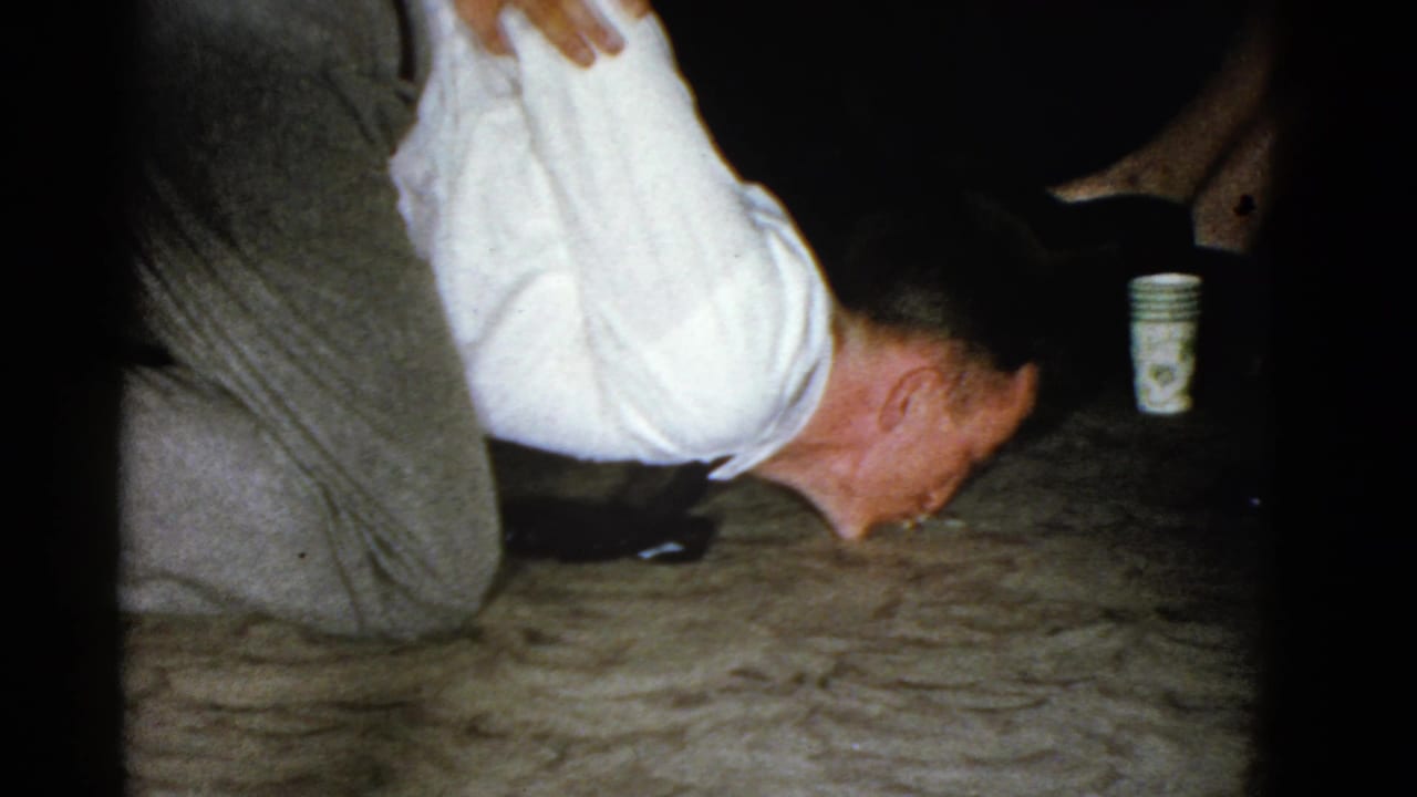 Man bends low over a carpet with his hands behind his back as a paper cup sits nearby.