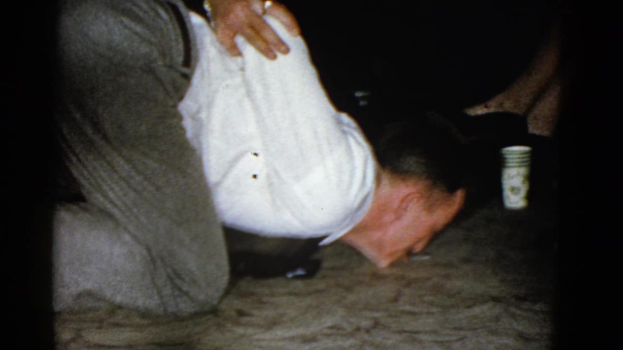 Man remains bent toward the carpet with a cup visible behind him in a dim room.