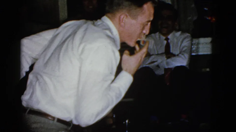 A man in a white shirt holds a small object or cigarette near his mouth as a seated man in a tie watches in the 1951 Boston interior.