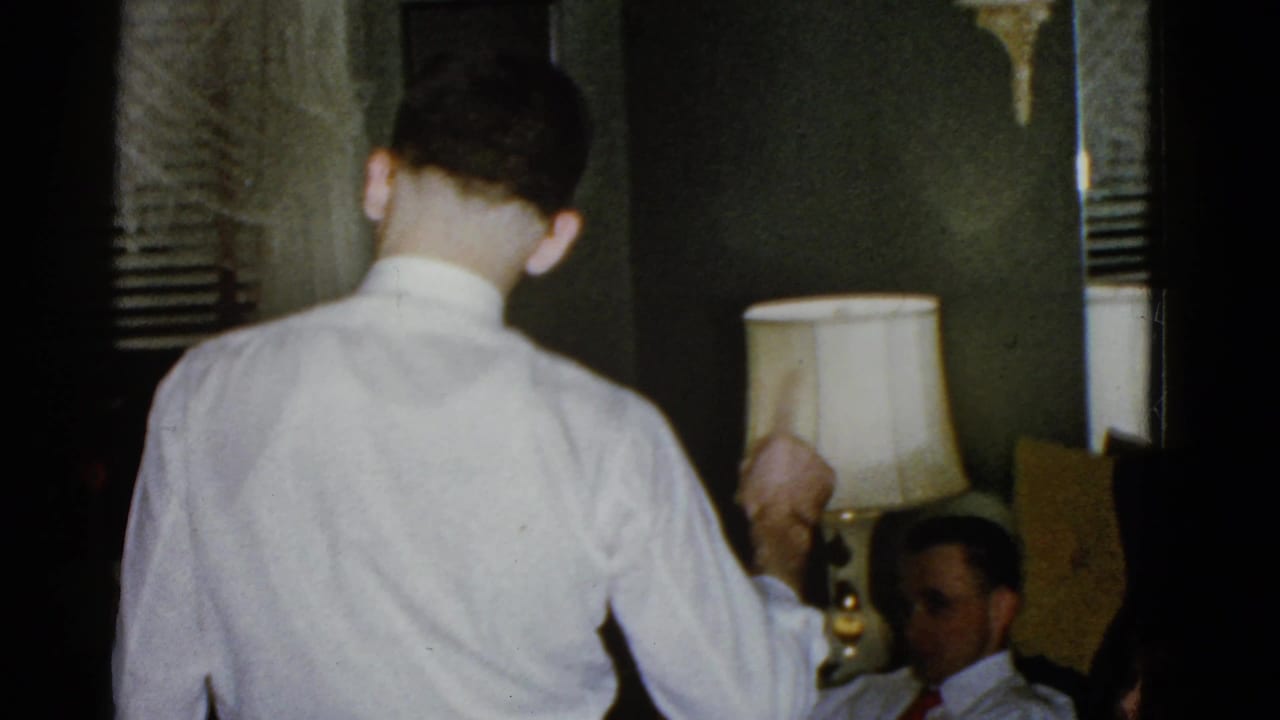 Man in a white dress shirt stands near a seated man in a dim room with a lamp.