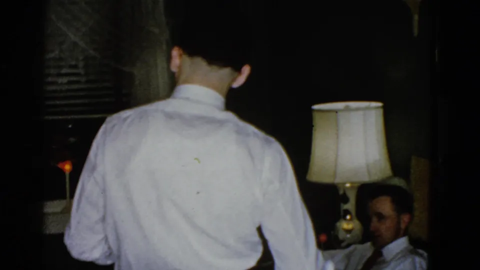 1951 Boston home-movie frame with a man seen from behind as a seated man watches from beside a lit table lamp.