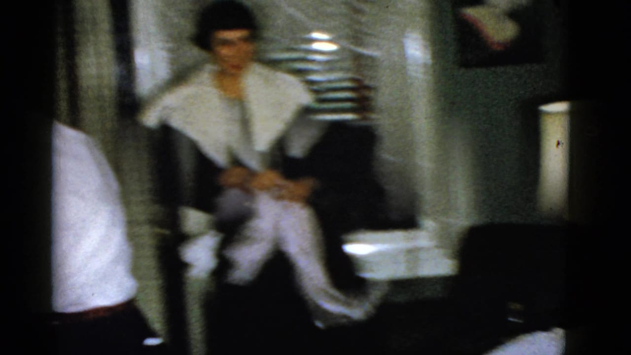 Dim room with a seated person near curtains and another figure partly visible at left.