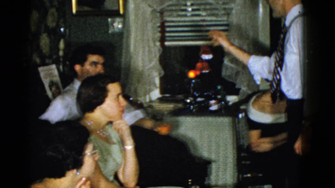 Standing man in a white shirt and tie gestures to seated adults during a 1951 Boston private gathering.