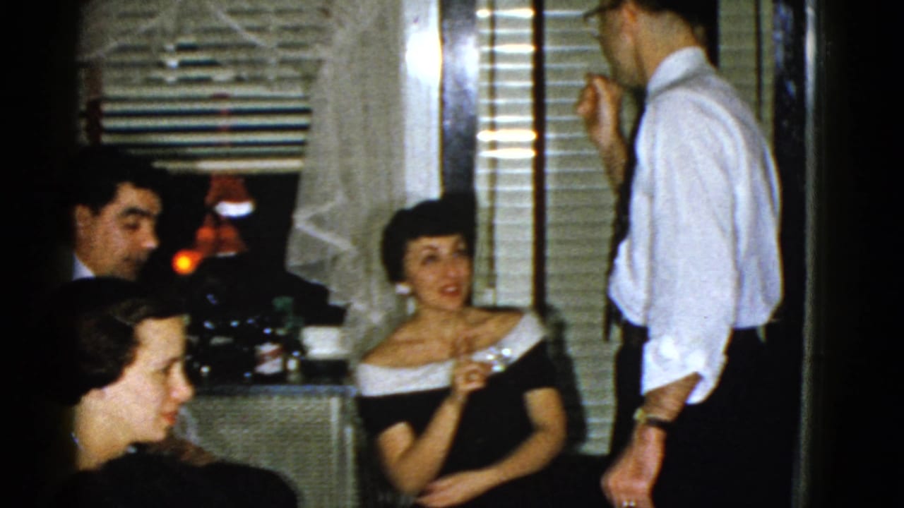 Adults in formal clothing converse in a dim room with window blinds and lace curtains.