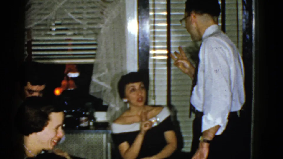 1951 Boston home-movie frame of seated adults reacting during conversation as a man stands near blinds and lace curtains.
