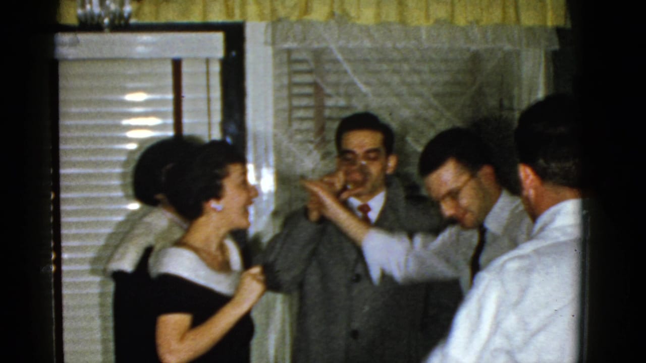 Adults in formal clothing gather near window blinds during an indoor reunion scene.