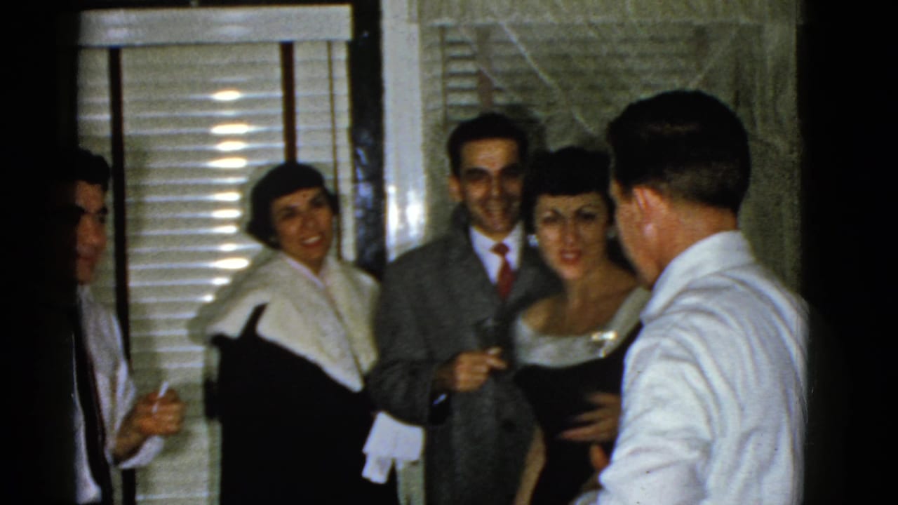 Adults in formal clothing gather indoors near window blinds and a doorway.