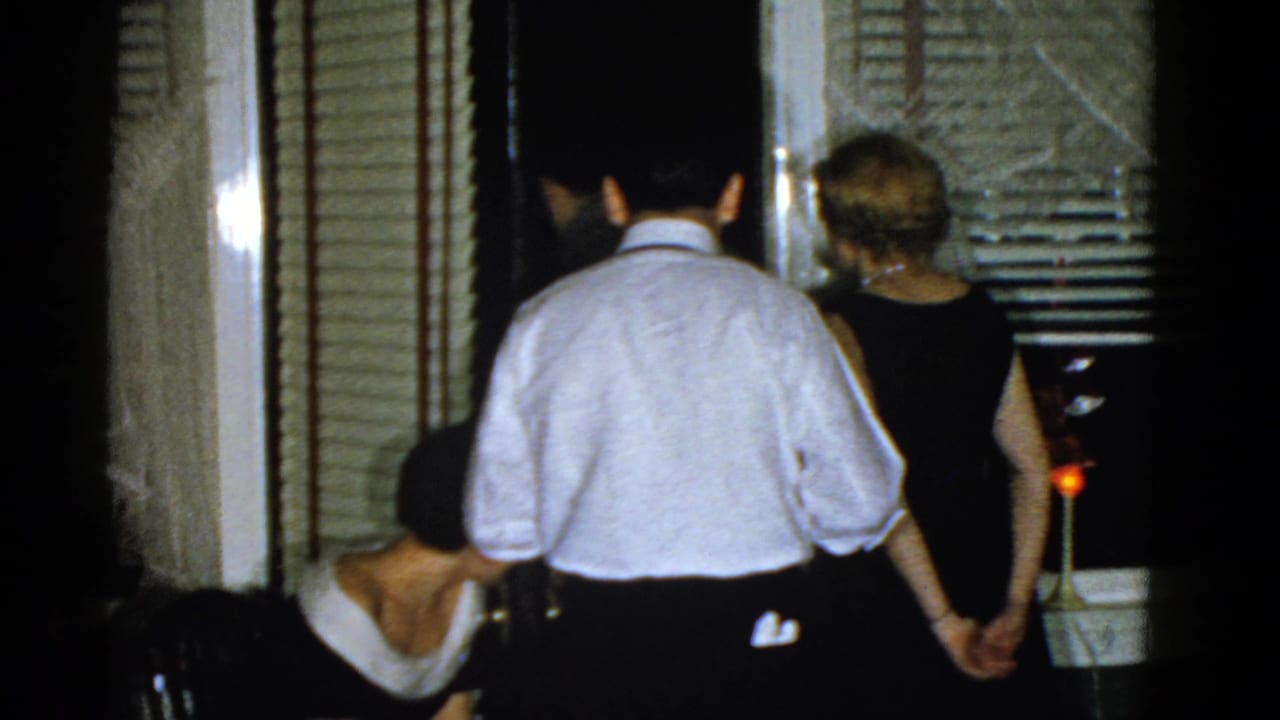 Adults in formal clothing stand and sit near a doorway with blinds and curtains.