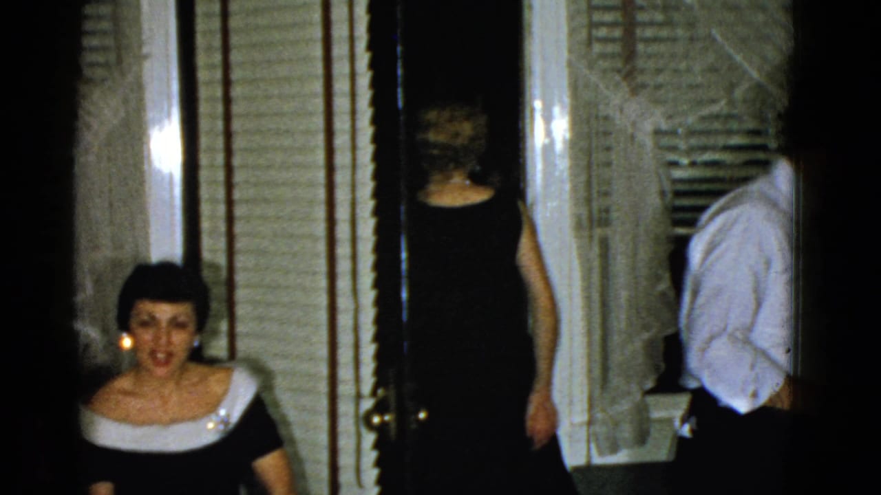 1951 Boston home-movie frame with a seated woman facing the camera as formally dressed adults move through the room.